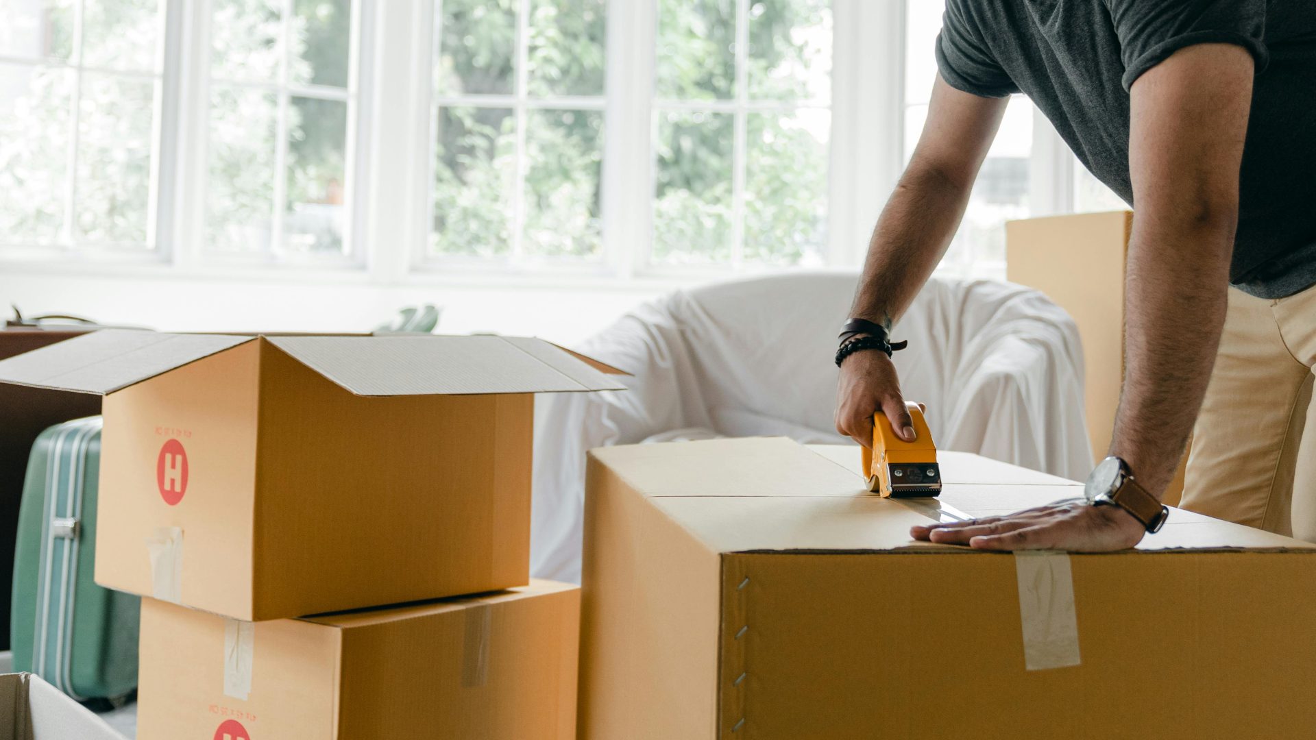 Hombre irreconocible con ropa informal empaca una caja de cartón usando un dispensador de cinta adhesiva para mudar artículos personales a un nuevo apartamento.