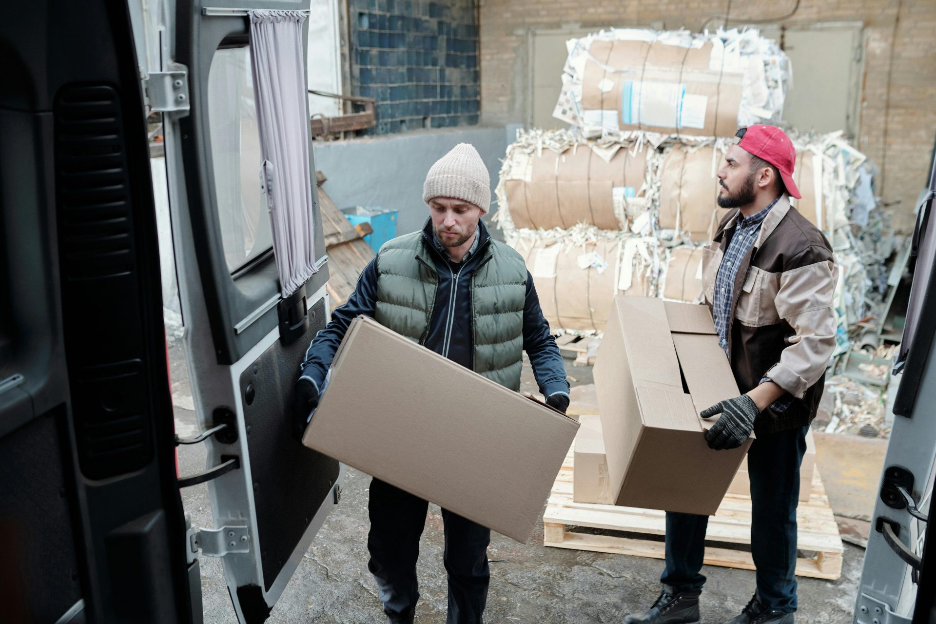 Dos trabajadores varones cargando cajas de cartón en un almacén, con paquetes de papel al fondo.