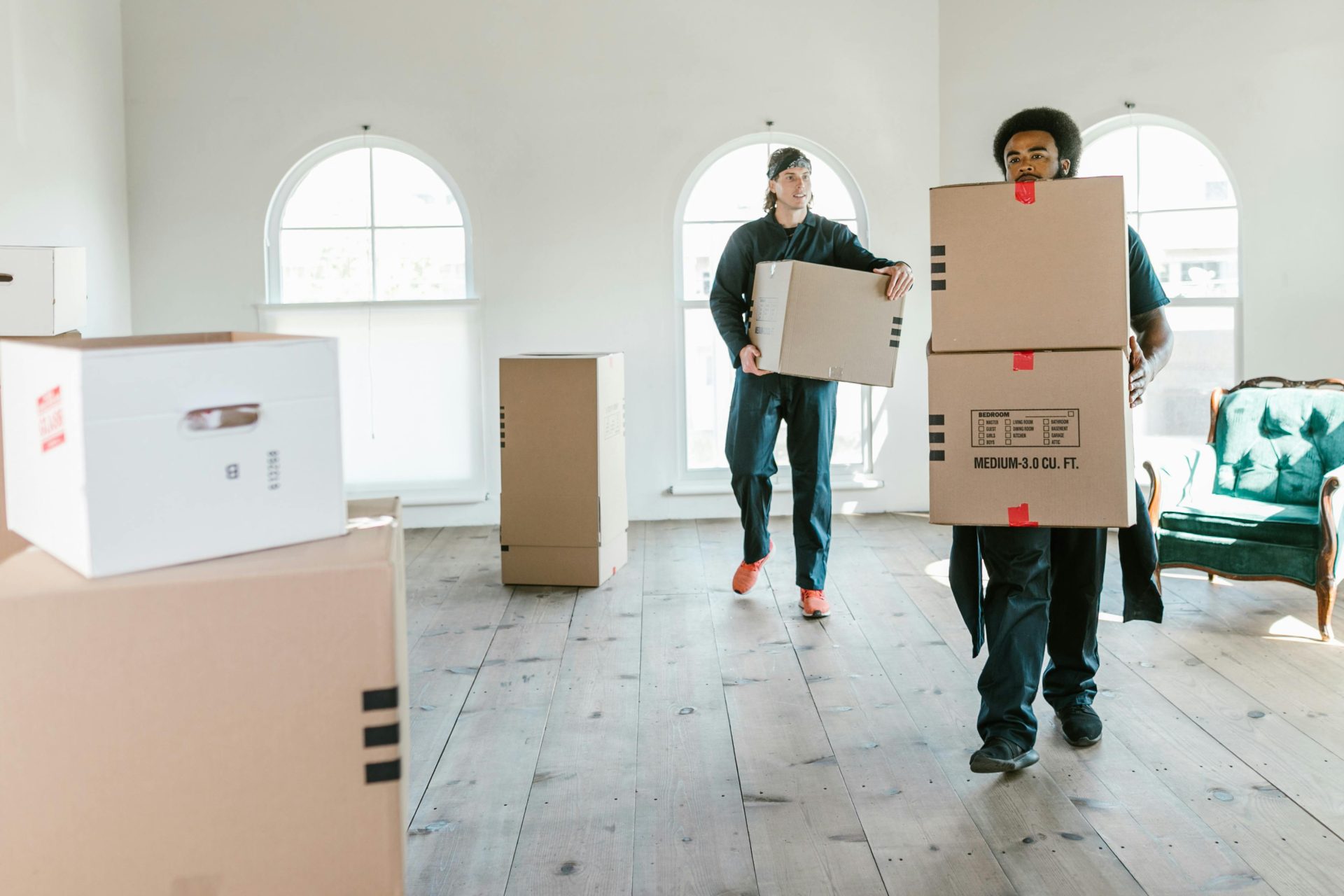Dos operarios de mudanzas cargando cajas en una habitación soleada, listos para ayudar con la reubicación.